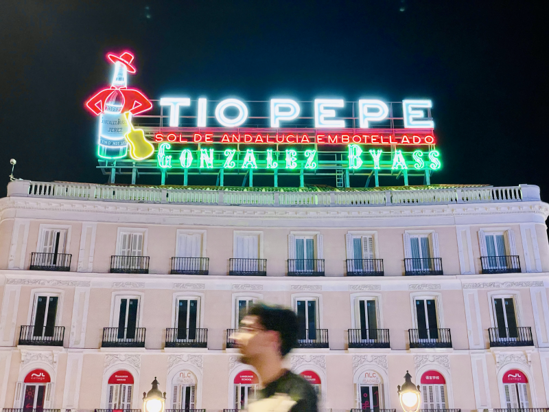 Night photography of a light pink building illuminated by colorful neon signs reading "TIO PEPE", "SOL DE ANDALUCIA EMBOTELLADO", and "GONZALEZ BYASS". A neon figure of a bottle wearing a red hat and holding a guitar is on the left. Blurry pedestrians and streetlights are in the foreground.